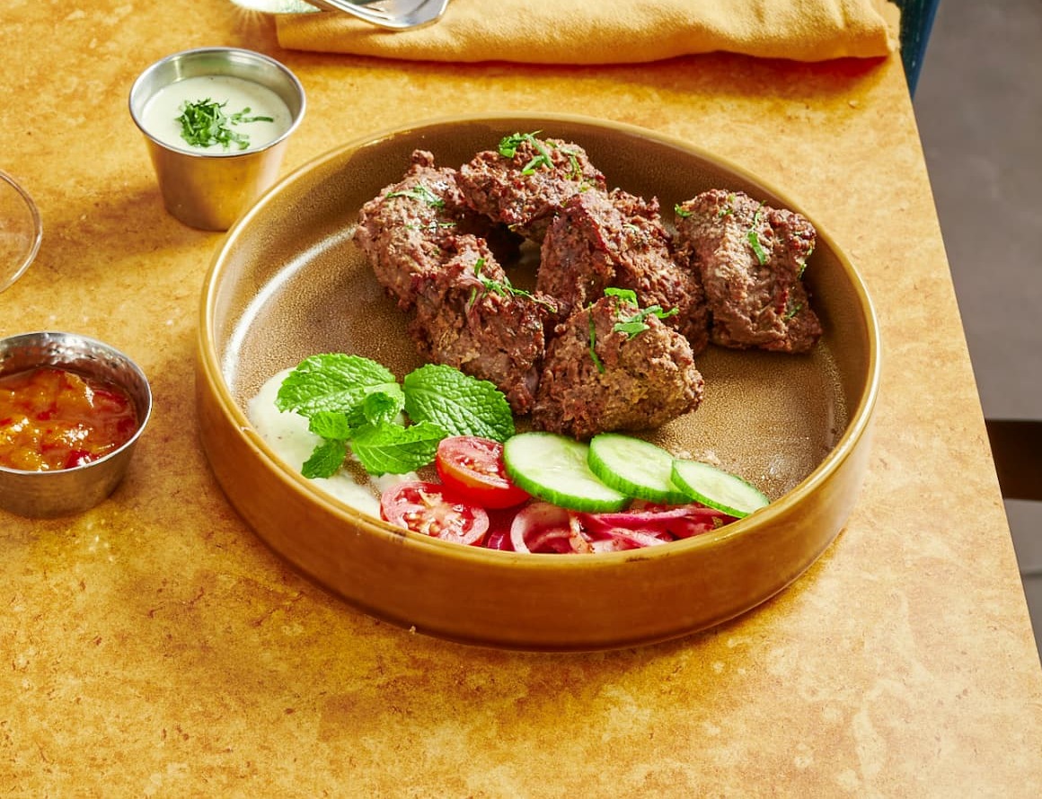 Original photograph of Peshawri lamb kebab served with cucumber, tomato and onion salad, mint, a creamy dip and a chilli relish on a golden table