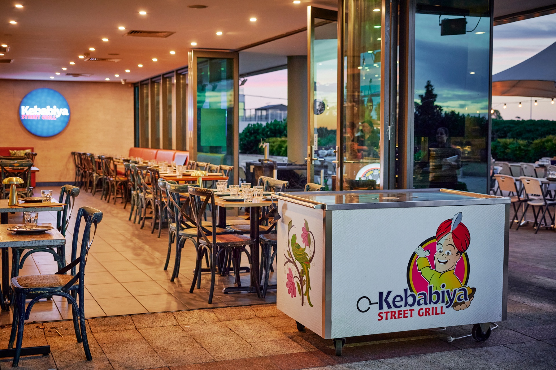 Kebabiya Street Grill dining room — long timber tables, cross-back chairs and the neon Kebabiya sign on the back wall