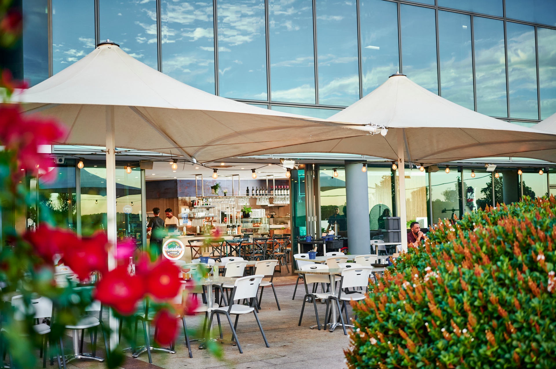 Kebabiya terrace under cream sail umbrellas with the Sky City glass façade behind and red flowers in the foreground